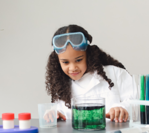 Young girl in lab coat with goggles watching science experiment.